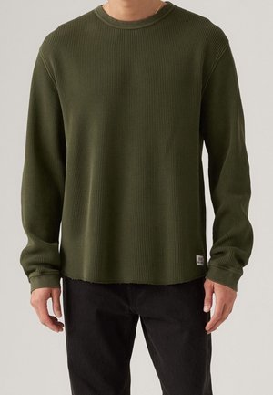 Mężczyzna ubrany w oliwkowo zieloną koszulę z długim rękawem o strukturze waflowej, ze ściągaczami na mankietach i małą metką z logo blisko dolnego brzegu, w połączeniu z czarnymi spodniami.