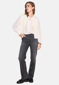 Un gilet di maglia crema sopra una leggera camicetta bianca con bottoni, abbinato a jeans a zampa d'elefante grigio scuro. Scarpe nere completano l'outfit.
