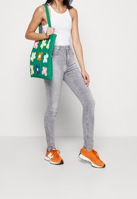 Tote bag verde con diseños de flores multicolores, combinada con jeans skinny grises y zapatillas naranjas, con acentos y cordones blancos.