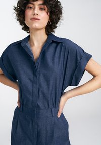 Granatowy kombinezon z krótkim rękawem, kołnierzykiem i paskiem w talii. Wykonany z materiału przypominającego denim o gładkiej teksturze.