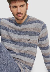 Suéter de punto a rayas en gris, azul y marrón con escote en V y un logo sutil en el pecho. Tejido suave con un acabado texturizado.