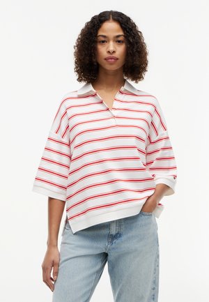 Kvinde med krøllet hår iført hvid polo med røde vandrette striber og lyseblå jeans, står med den ene hånd i lommen.