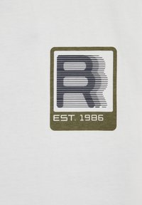 Balta medvilnė marškinėliai su spaudiniu: dėžutės logo juodais kontūrais ir alyvuogių žalia apvadu, po juo užrašas „EST. 1986“. 