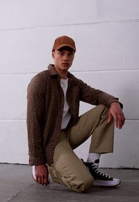 Braun kariertes Hemd über einem weißen Hemd, beige Hose und schwarze Sneakers mit weißen Sohlen. Die Person kniet vor einem neutralen Hintergrund.