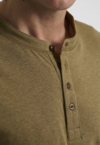 Camisa henley de manga corta en verde oliva, hecha de algodón suave, con cuello redondo, tres botones en el cuello y una textura sutil.
