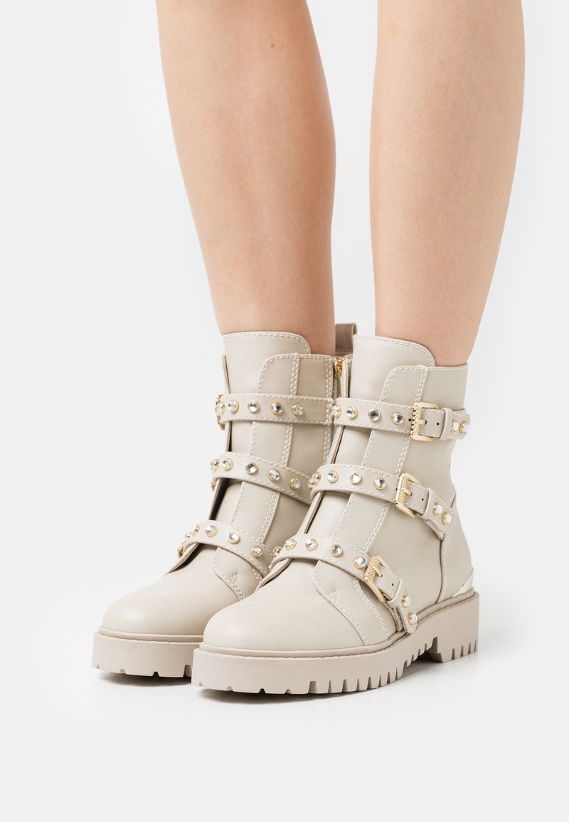 Bottines beiges à semelles épaisses, avec trois brides cloutées munies de boucles dorées, portées sur des jambes nues sur un fond blanc uni.