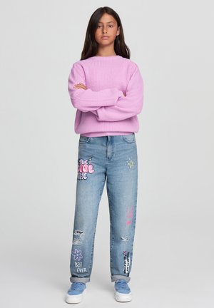 Maglione rosa lavorato a maglia con un motivo testurizzato, abbinato a jeans chiari sfilacciati che presentano stampe grafiche colorate e una vestibilità comoda.
