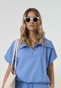 Gaiši zils ceturtā daļa ar rāvējslēdzēju tops ar platām piedurknēm un reljefām burtām. Modelis valkā baltas kaķu acs saulesbrilles un tur gaiši brūnu somu.