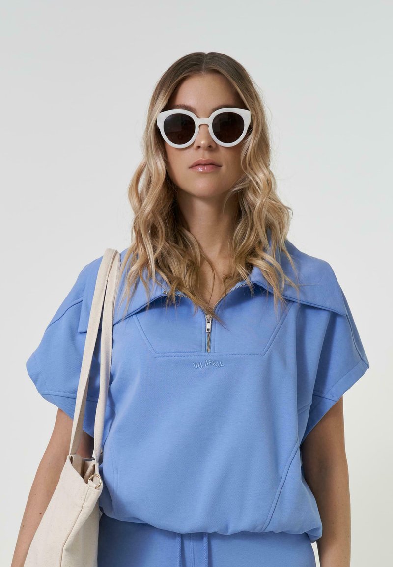 Gaiši zils ceturtā daļa ar rāvējslēdzēju tops ar platām piedurknēm un reljefām burtām. Modelis valkā baltas kaķu acs saulesbrilles un tur gaiši brūnu somu.