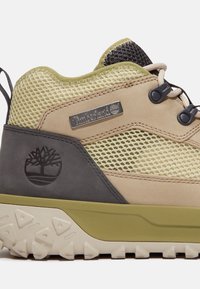 Brun och olivfärgad vandringsko med meshuppersidan, gummisulor, Timberland-logotyp och förstärkta detaljer för hållbarhet och stöd.