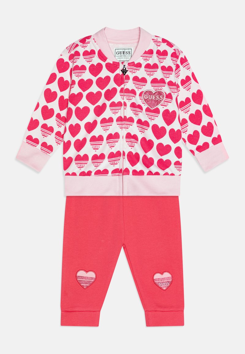 Giacca con zip a cuore rosa con motivo a cuori e patch con logo, abbinata a pantaloni rosa uni con dettagli applicati a forma di cuore sulle ginocchia.