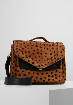 Sacoche tachetée marron et noire avec rabat en cuir noir et bandoulière réglable sur une surface d'exposition blanche.