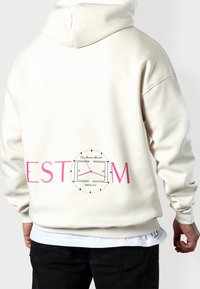 Krémová mikina s kapucí a šňůrkou, na zádech růžový nápis "EST" a grafika hodinek. Měkká látka s žebrovanými manžetami a lemem.