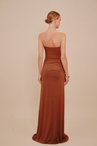 Bunba kötött hajú nő egy vállnélküli, földig érő barna ruhában áll egy bézs háttér előtt, háttal a kamerának.