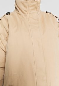 Beżowa, lekka kurtka z wysokim kołnierzem, z naramiennikami i widocznym przeszyciem. Teksturowana tkanina o gładkim wykończeniu.