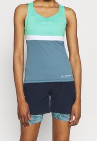Persona che indossa una maglietta sportiva senza maniche a blocchi di colore in turchese, bianco e blu, con pantaloni corti blu navy e pantaloni da bici blu-verdi a fantasia sotto.
