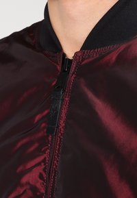 Bordowa kurtka bomberka o gładkim, błyszczącym wykończeniu; posiada czarny prążkowany kołnierz oraz zamek błyskawiczny z fakturowanym akcentem w pobliżu suwaka.