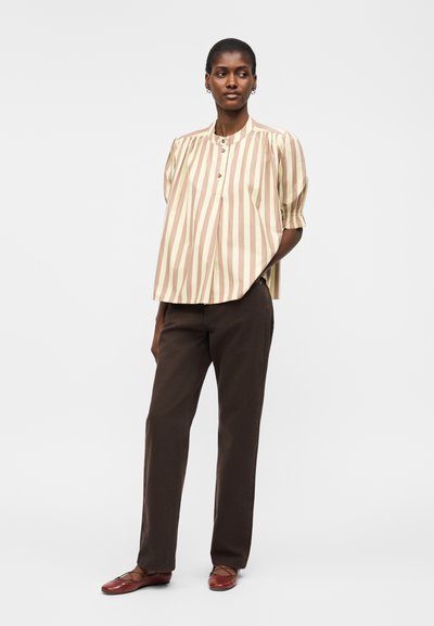 Γυναίκα όρθια με τα χέρια στις τσέπες, φοράει μπλούζα με κρεμ και καφέ κάθετες ρίγες και κουμπιά, σκούρο καφέ παντελόνι και μπορντό φλατ παπούτσια.