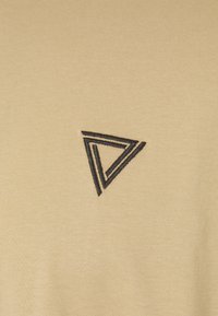 Camiseta beige claro con un diseño geométrico de triángulo negro bordado en el lado izquierdo, hecha de un tejido suave y liso.