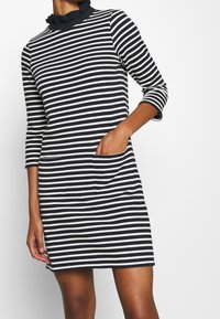 Raidallinen mekko korkealla kauluksella ja rypyttömällä kauluksella, jossa on kapea malli, pitkä hihat ja kaksi etutaskua. Värit: tummansininen ja kerma.