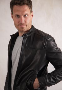 Black leather jacket with a stand collar, zip closure, and side pockets; worn over a gray t-shirt, featuring smooth, glossy texture.