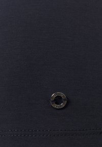 Schwarzer Stretchstoff mit glatter Textur, ausgestattet mit einem kleinen Metallösen und dem Text "OLIVER" sowie einer sichtbaren Nahtdetail.