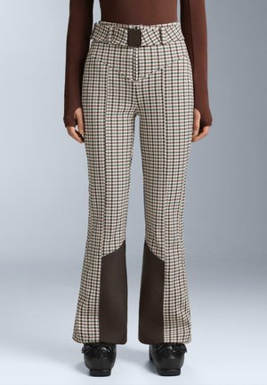 Donna che indossa pantaloni a vita alta in houndstooth con pannelli marroni alle caviglie, abbinati a scarpe platform nere e un top a maniche lunghe marrone.