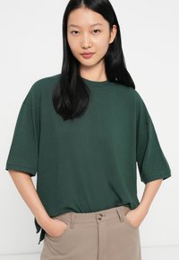 Fiatal nő, aki hosszú fekete hajat visel, sötétzöld, túlméretezett, rövid ujjú pólót és bézs nadrágot hord, kezét a zsebében tartva.