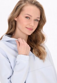 Világoskék, cipzáros kapucnis pulóver, magas gallérral, puha anyagból és bordás mandzsettával, laza szabással és minimális varrási részletekkel.