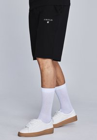 Černé sportovní šortky s elastickým pasem, s bílým logem na levé přední straně. Vytvořené s bílými ponožkami a bílými teniskami.