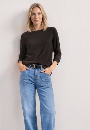 Kvinde med blondt hår iført en sort langærmet top stukket ned i lyseblå højtsiddende jeans med sort bælte, stående mod en ensfarvet væg.