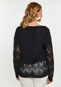 Schwarzer gestrickter Pullover mit einem Zickzackmuster, transparenten Ärmeln und einem kontrastierenden weißen Zickzackdesign am Saum. Weicher Stoff und lockere Passform.