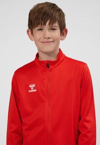 Giacca rossa con zip, realizzata in tessuto liscio, con collo alto e il logo bianco "Hummel" sul lato sinistro del petto. Sfondo neutro.