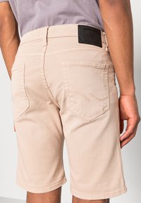 Beige farkkushortsit suoran leikkauksen omaavina, varustettuna takataskuilla, ompelutiedoilla ja mustalla tarralla vyötäröllä.