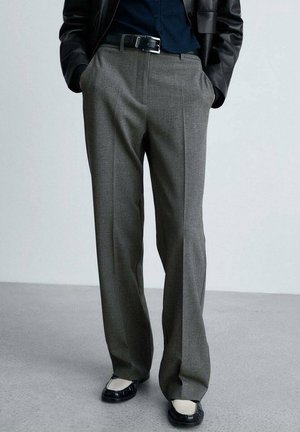 Personne portant un pantalon gris droit, une ceinture noire, une veste en cuir noire, une chemise bleu marine et des mocassins noirs et blancs, debout les mains dans les poches.