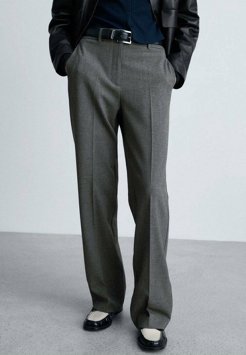 Personne portant un pantalon gris droit, une ceinture noire, une veste en cuir noire, une chemise bleu marine et des mocassins noirs et blancs, debout les mains dans les poches.