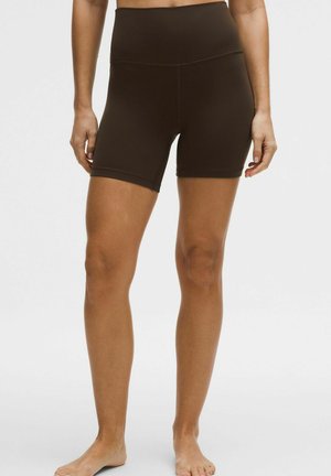 Žena v tmavohnedých priliehavých cyklistických šortkách s vysokým pásom stojaca bosá na bielom pozadí.