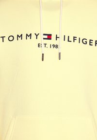 Felpa gialla con tasca anteriore, con il logo "TOMMY HILFIGER" in blu scuro e una piccola grafica di bandiera rossa e bianca.