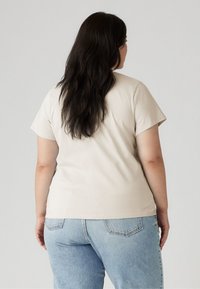 Lys beige kortermet t-skjorte med rund hals, kombinert med høyde i livet blå denimbukser, vist fra baksiden.