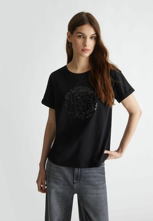Maglietta nera in cotone con un design rotondo di paillettes al centro, maniche corte e una vestibilità rilassata. Abbinata a jeans in denim grigi.