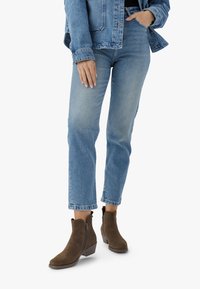 Femeie îmbrăcată în blugi albaștri deschiși, o jachetă din denim și ghete din suede maro cu toc mic, stând cu o mână în buzunar.