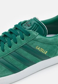 Zöld velúr sportcipő három sötétebb zöld csíkkal és arany "Gazelle" felirattal. Párnázott bokarésze és fehér gumitalpa van.