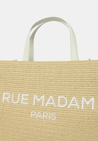 Borsa tote in paglia intrecciata beige con testo ricamato bianco "RUE MADAM PARIS." Presenta due manici in ecopelle bianca. Forma rettangolare.