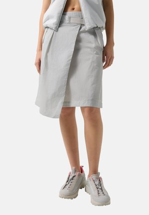 Kobieta ubrana w jasnoszarą spódnicę z nakładanym przednim panelem, bezrękawnik i masywne białe sneakersy z różowymi akcentami.