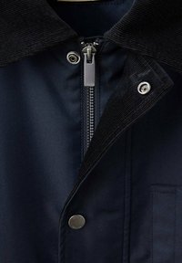 Gros plan d'une veste bleu marine foncé avec un col en velours côtelé, une fermeture éclair visible et des boutons-pression métalliques à l'avant.