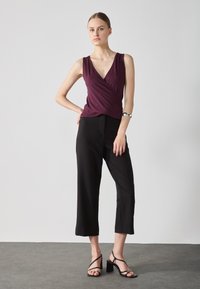 Bordeaux mouwloze top met een V-hals en wikkelontwerp, gecombineerd met zwarte cropped broek en sandalen met blokhak en bandjes. Effen textuur.
