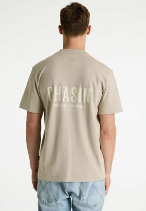 Man met kort bruin haar, gekleed in een beige T-shirt met de tekst "CHASIN' AMS BASED - C.COMMUNITY" op de achterkant, en lichtblauwe spijkerbroek.