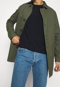 Olivgrön, lätt kappa med krage och sex svarta knappar, bärs över en mörk tröja och blå jeans. Slät textur, avslappnad design.