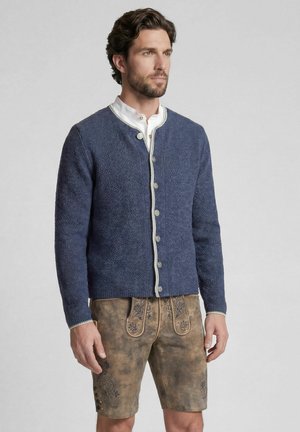 Mann trägt traditionelle braune bestickte Lederhosen und eine marineblaue geknöpfte Strickjacke über einem weißen Hemd, steht vor grauem Hintergrund.