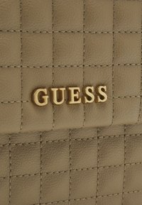 Sac en simili cuir matelassé beige avec un logo "GUESS" en doré et des détails de couture contrastants. Forme rectangulaire compacte.